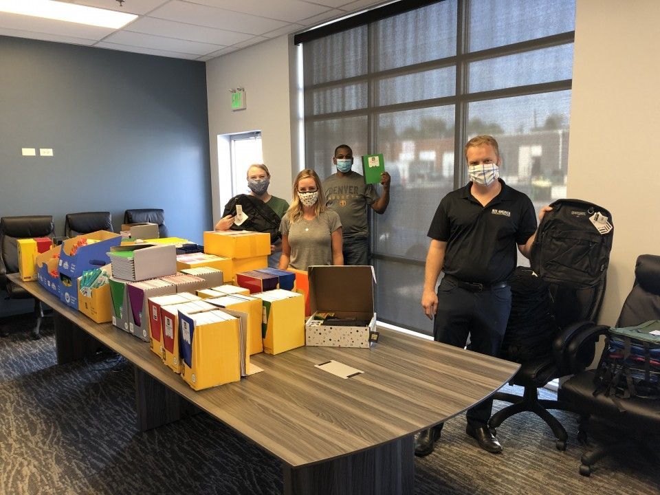 Five people wearing masks stand behind a table covered with boxes and folders in an office setting.