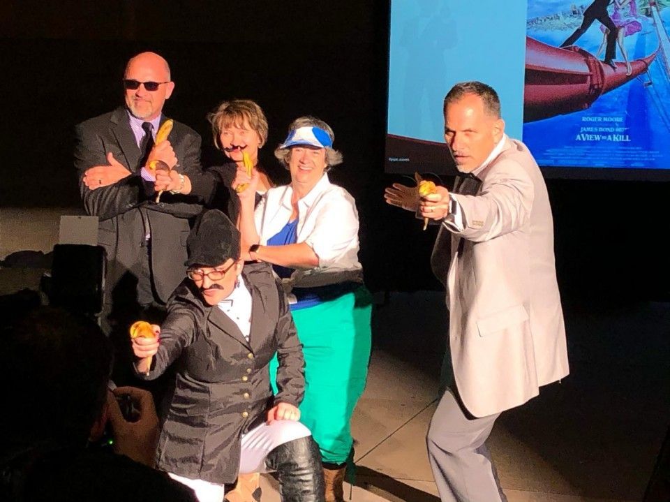 Group of people posing with bananas, some aiming them. A movie screen in the background.