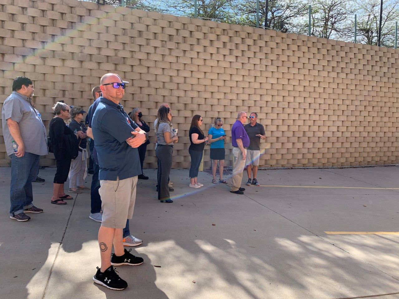 Group of people standing in front of a textured retaining wall outdoors, listening to a speaker.