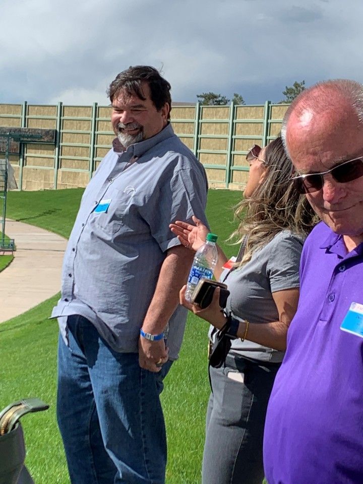 Three people outside on a sunny day; man in blue shirt smiles, woman gestures with bottle, man in sunglasses looks down.