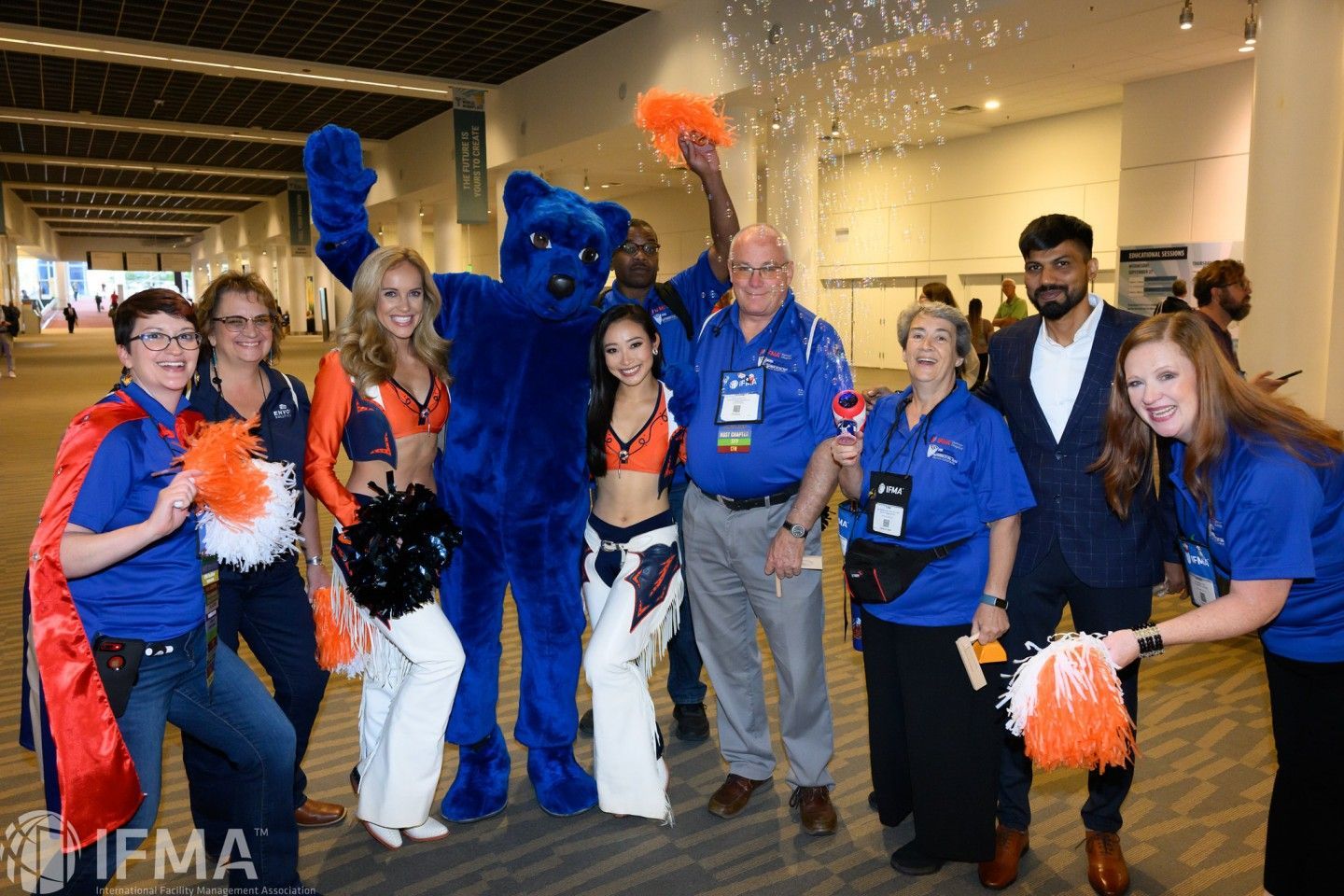 A group of people, including a mascot, cheerleaders, and staff, pose in a large hallway.