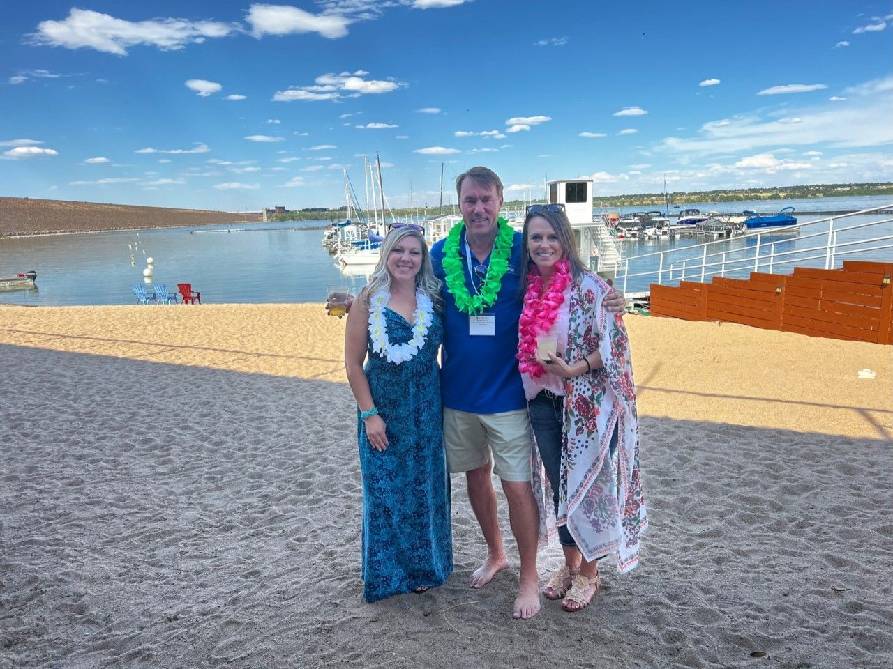 Three people with leis on a beach with a marina in the background under a blue sky.
