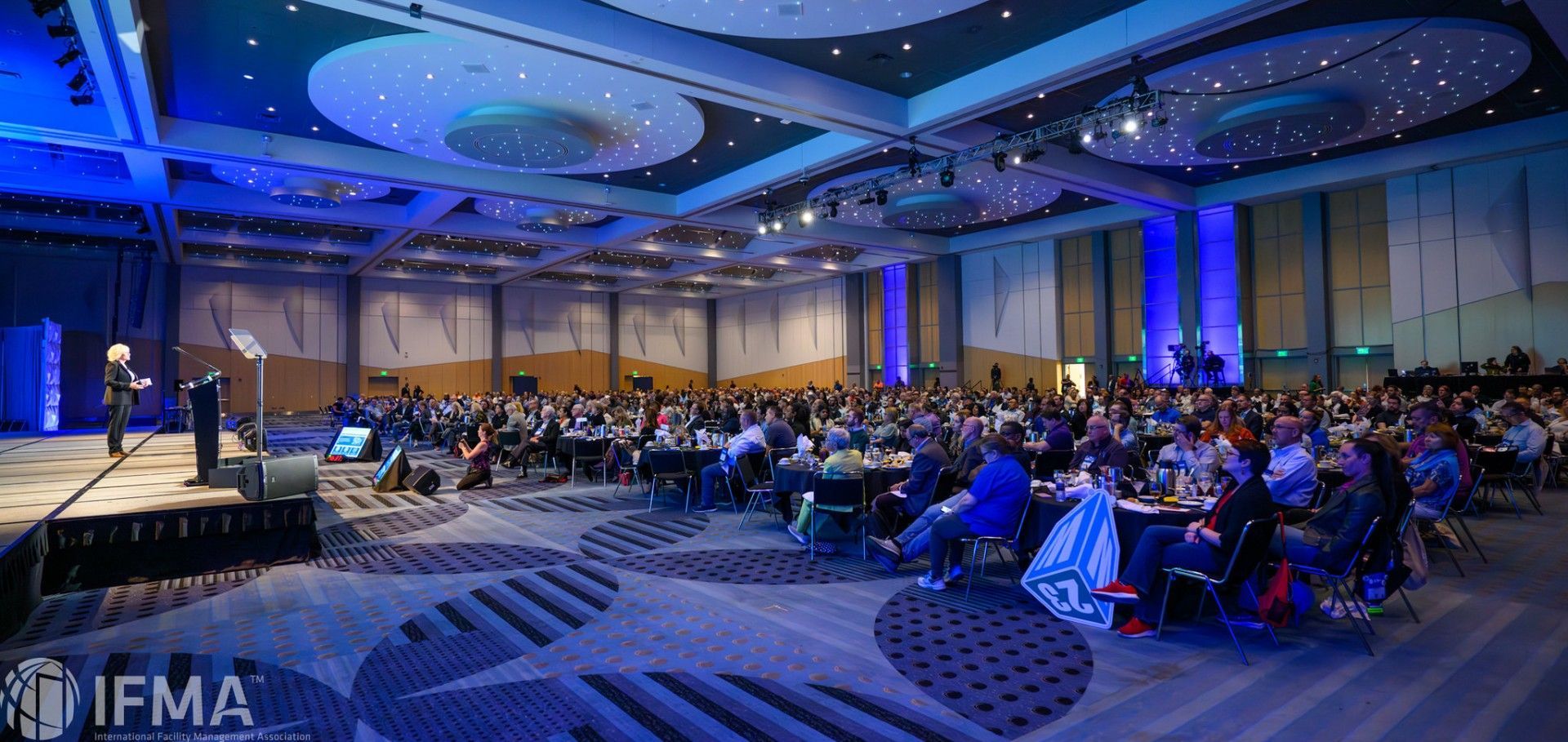 Large conference hall with a speaker on a stage addressing a seated audience, under blue lighting.