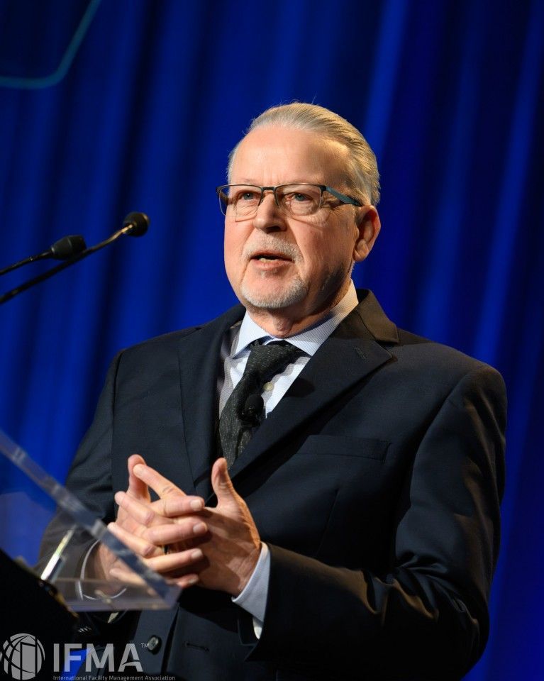 Man in dark suit speaks at a podium; blue backdrop.
