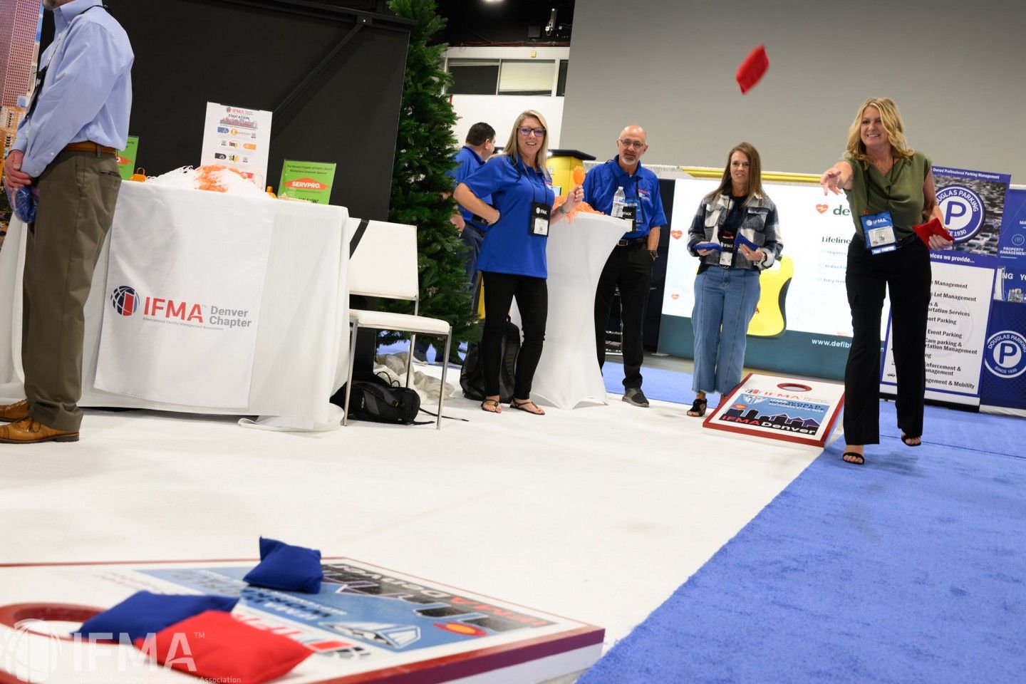 People playing bean bag toss at an event. Woman throws bean bag; others watch. Indoor setting.