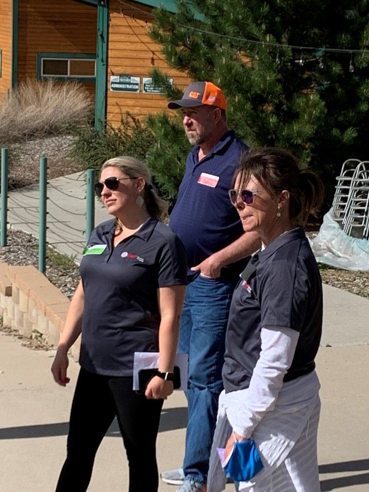 Three people outdoors: two women and a man. All wearing similar dark shirts. Man in orange hat. Sunny day.