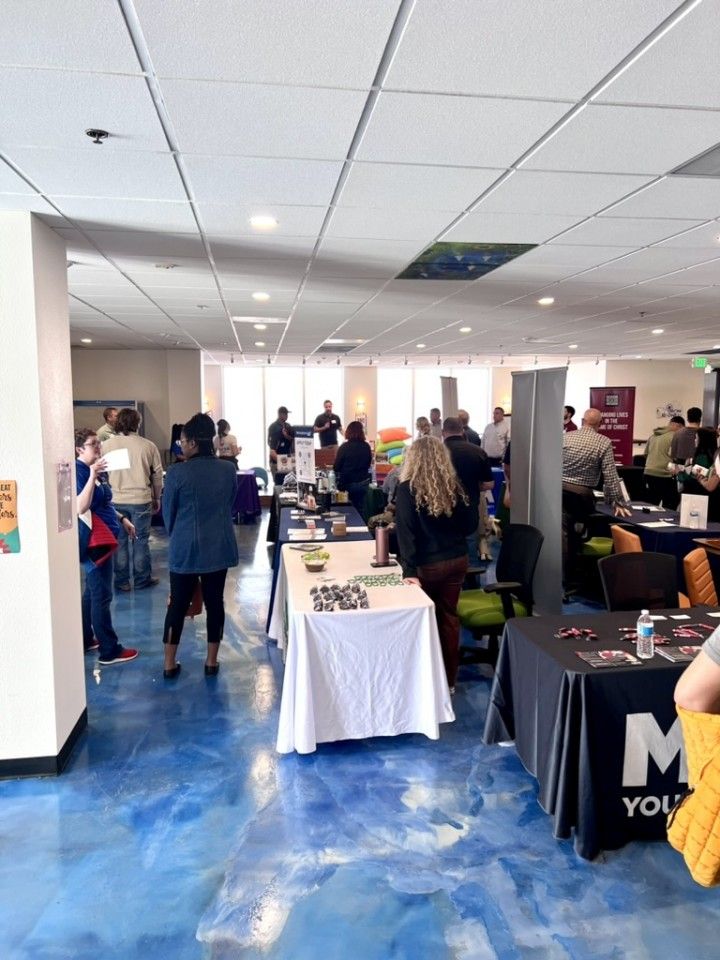People at a career fair, standing and looking at booths with tables. Blue floor, white tables and ceiling.
