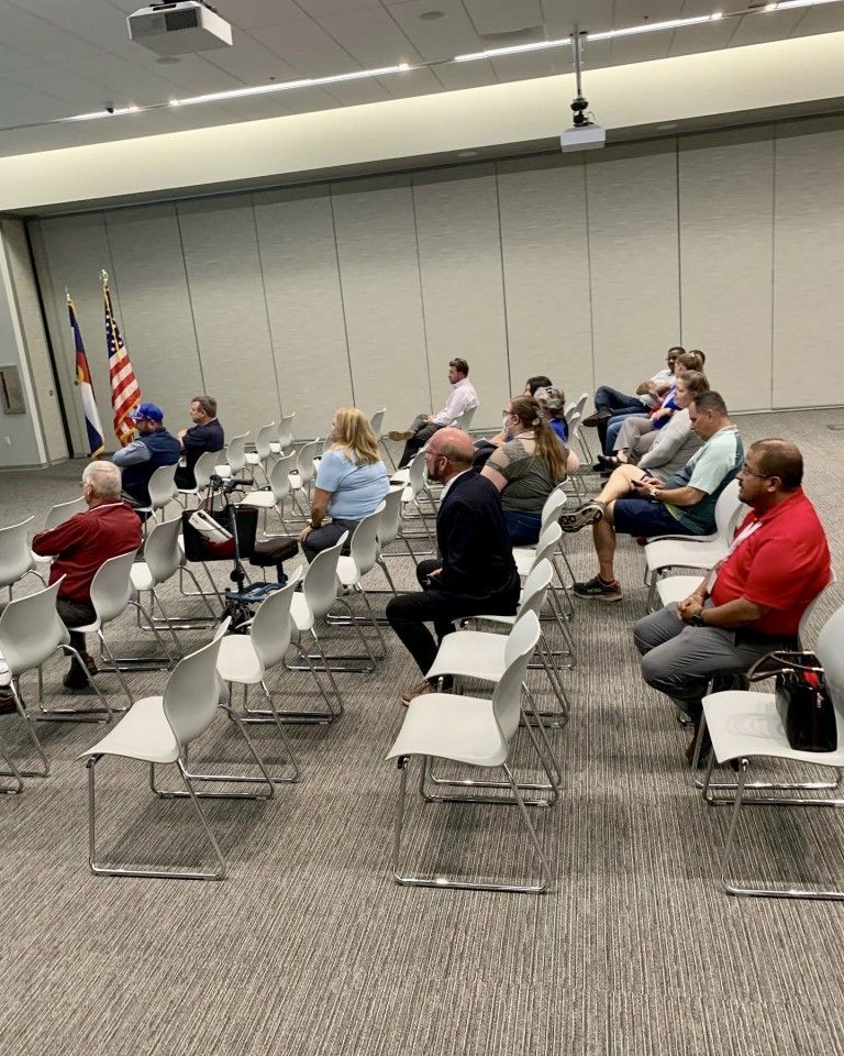Audience sitting in a meeting room, facing forward. American and Colorado flags. Chairs, light-colored walls.