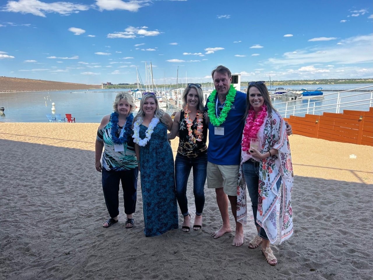 Five people, wearing leis, pose on a beach. A marina is visible in the background under a blue sky.