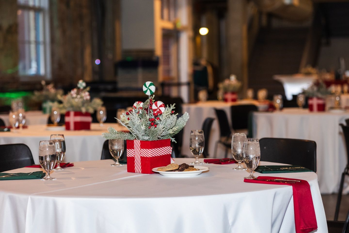 Round tables set for a holiday event, featuring red and green centerpieces and decorations.