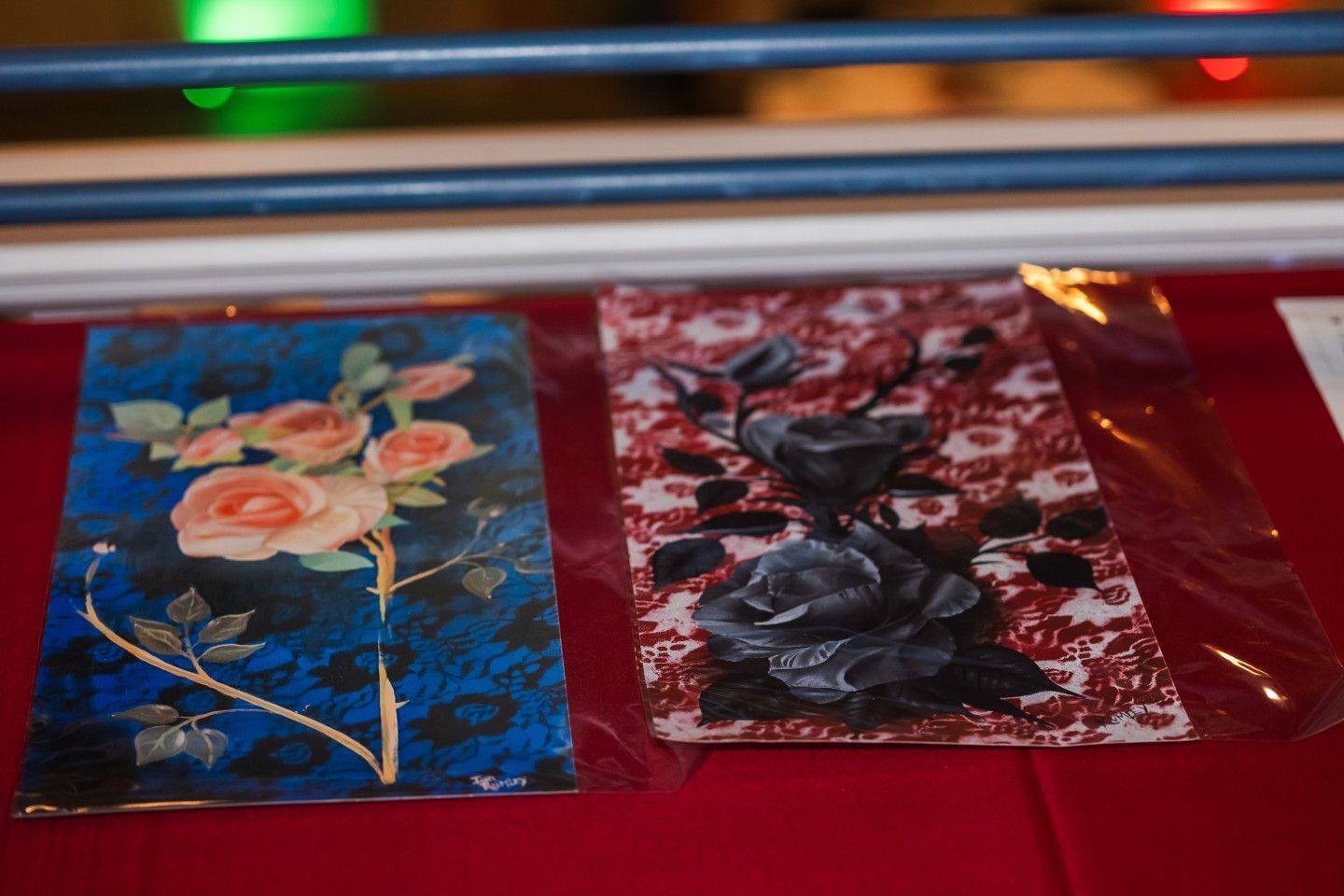 Two floral paintings: roses on blue and black roses on red patterned background, displayed on a red surface.