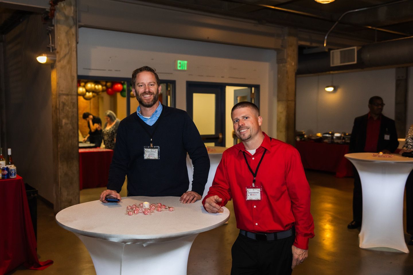Two men at an event, standing by a table with candy. One wears a red shirt, the other a sweater.