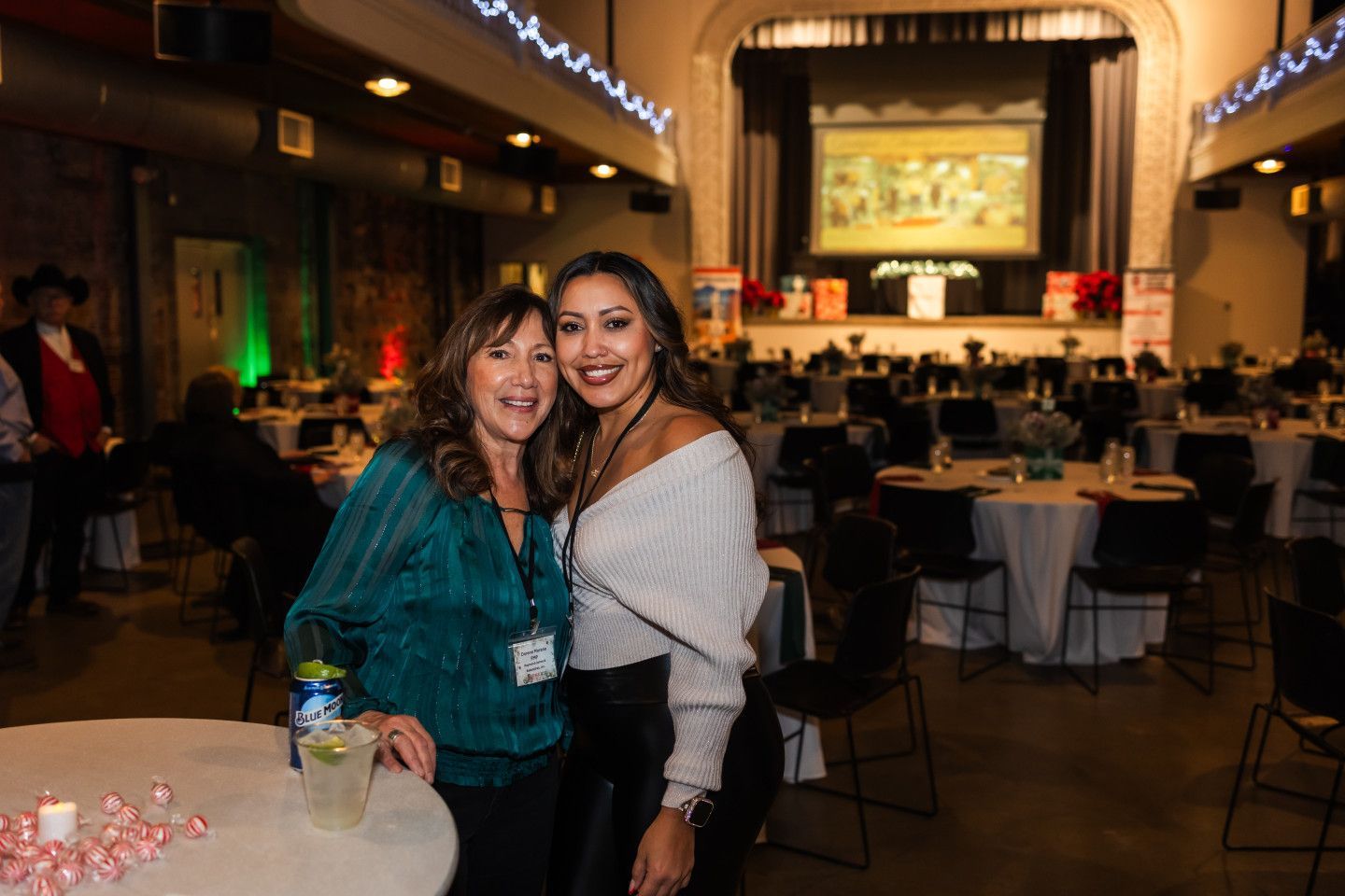 Two women smile, pose together at a banquet hall. Tables, chairs, and a screen in the background.