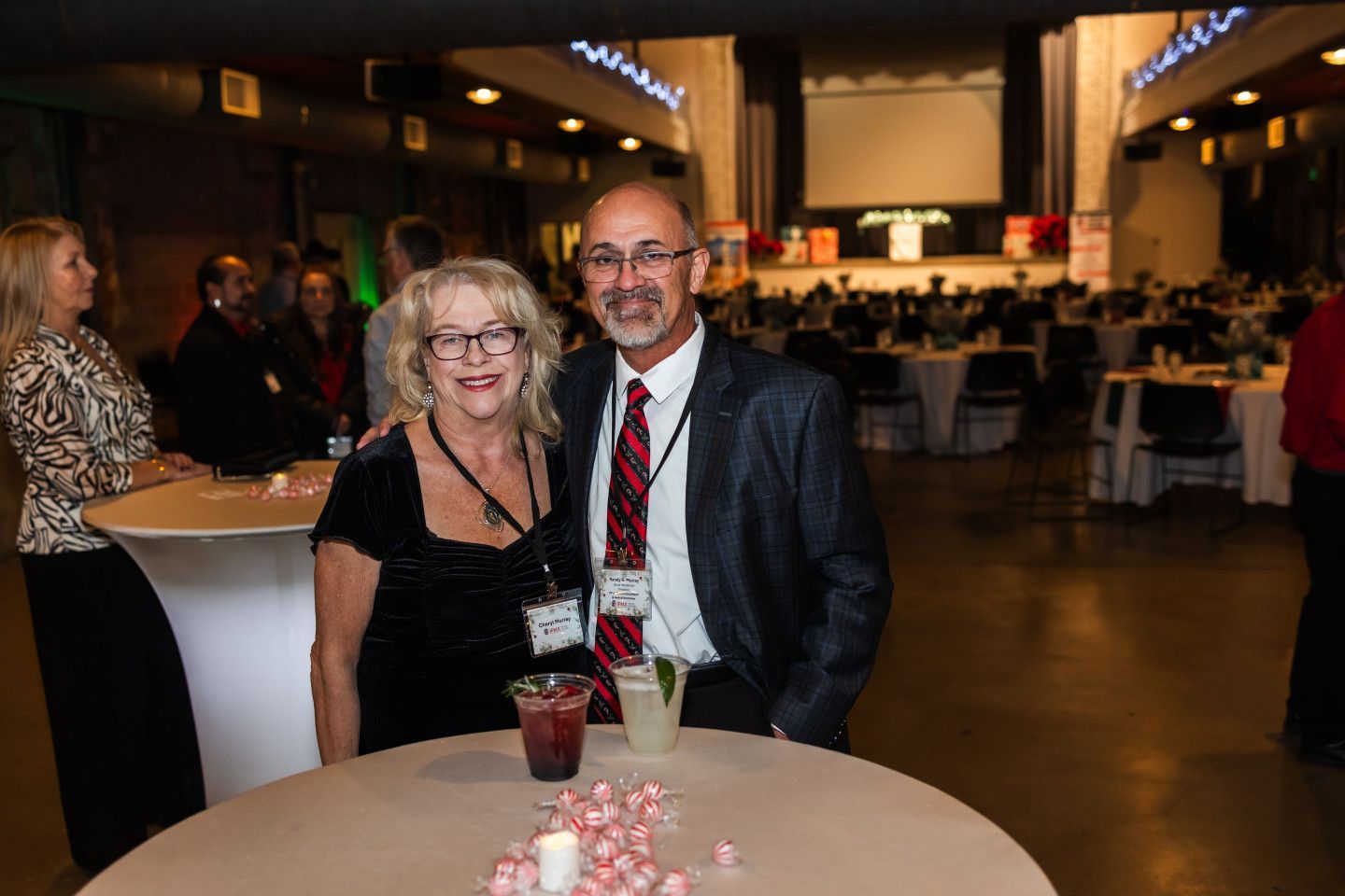 Couple at event, posing near cocktail table with drinks. Event space in background.
