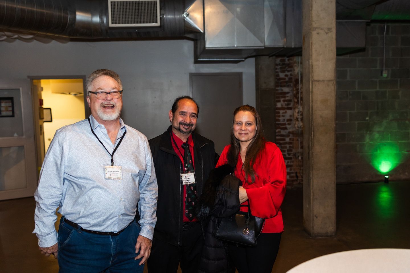 Three people smile, indoors. Man in blue shirt laughs. Man in black coat. Woman in red shirt. Green light on wall.
