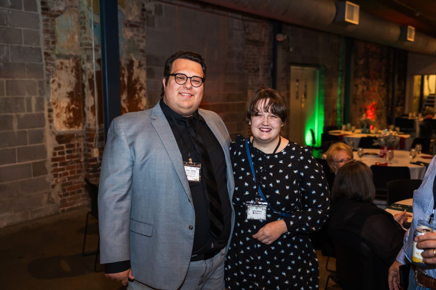 Two people pose for a photo in a brick-walled room; man in a grey suit, woman in a black patterned dress.