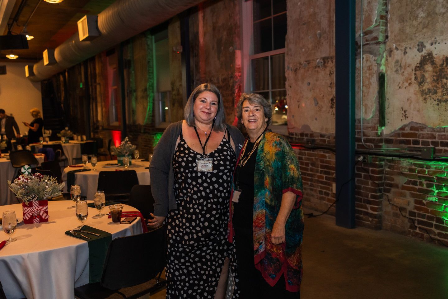 Two women pose at a well-lit event. One wears a black dress, the other a patterned shawl. Tables are set in the background.