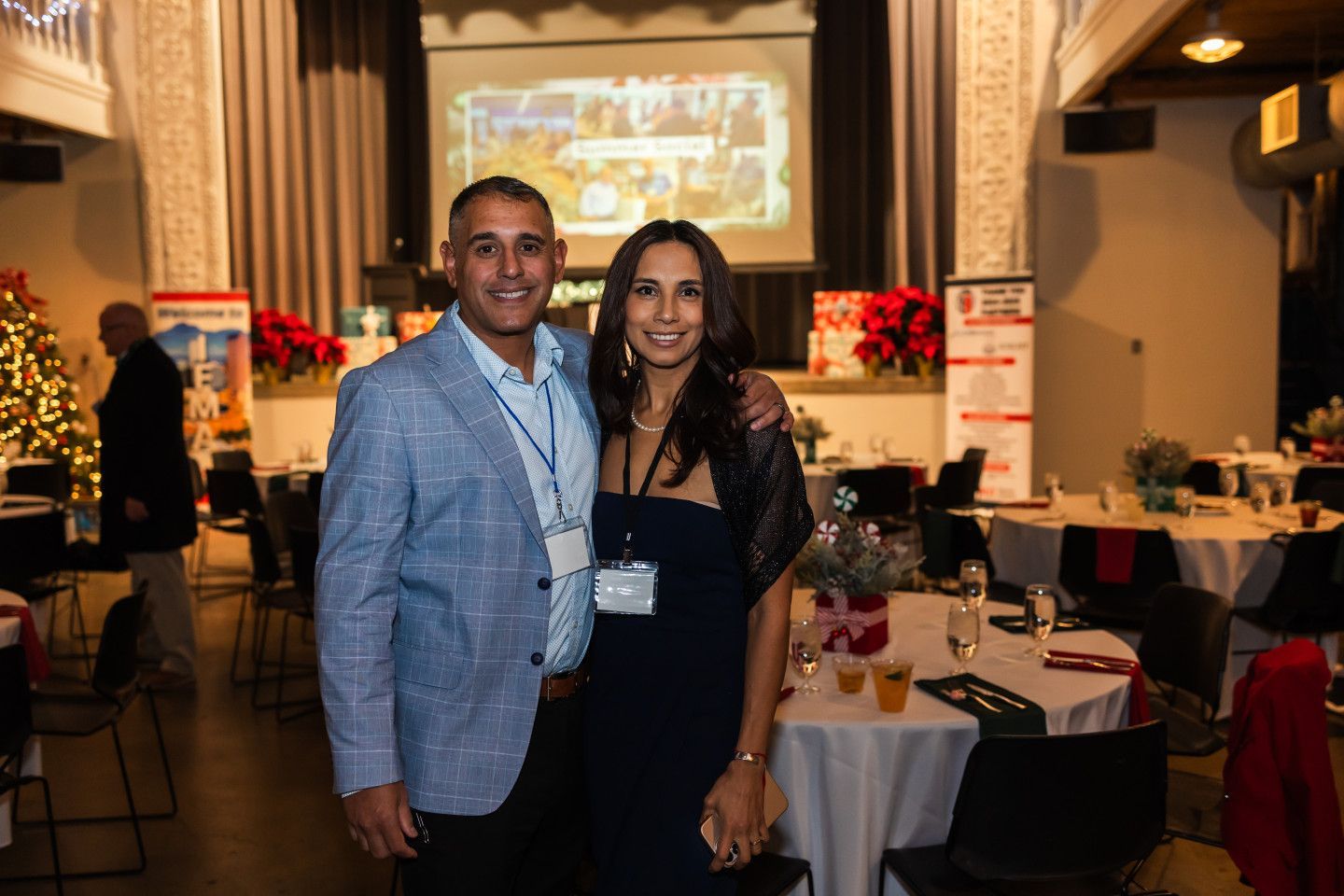 A man and a woman smile, posing together at an event. They stand near tables with decorations.