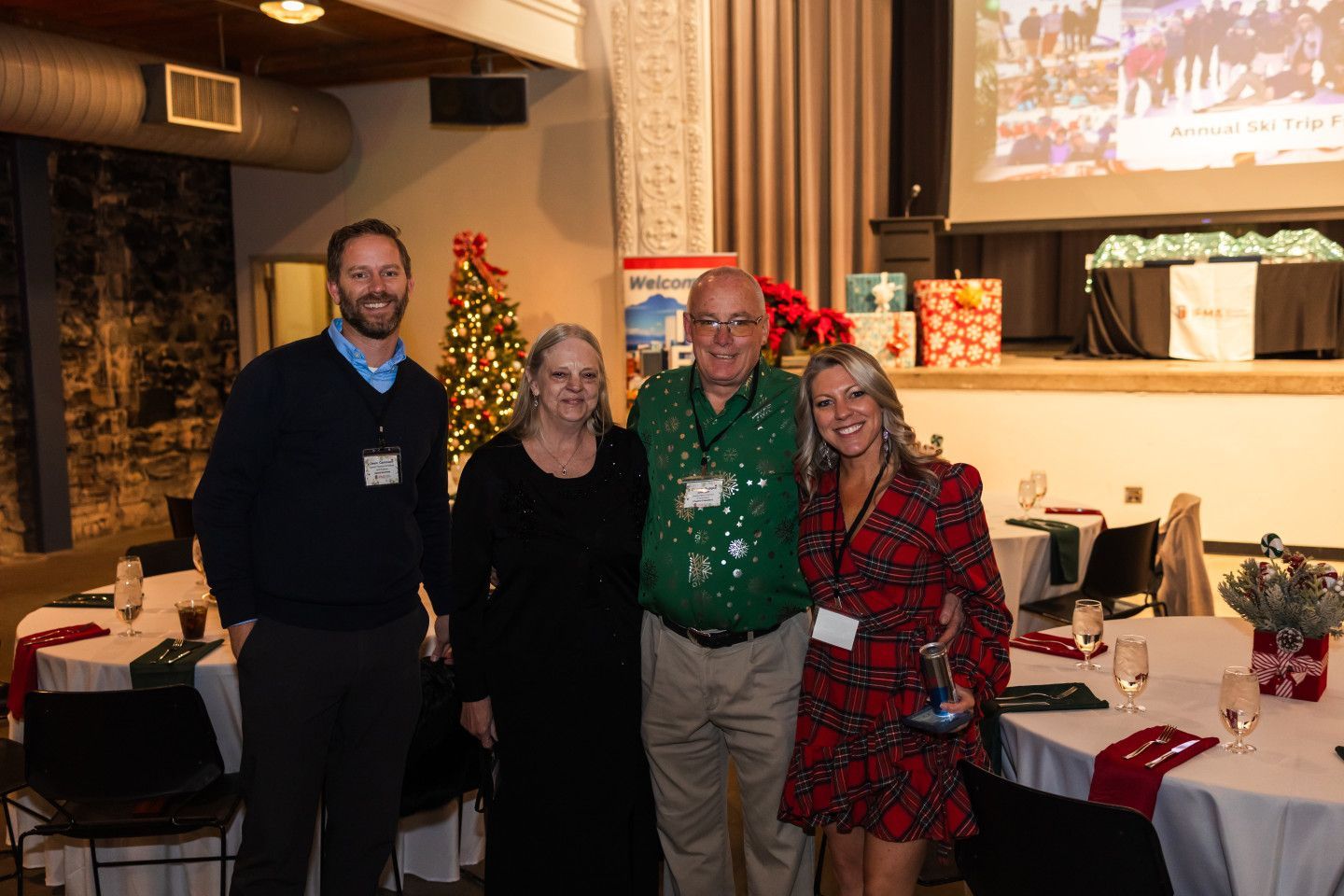 Four people pose at a holiday event, next to a Christmas tree and decorated tables.