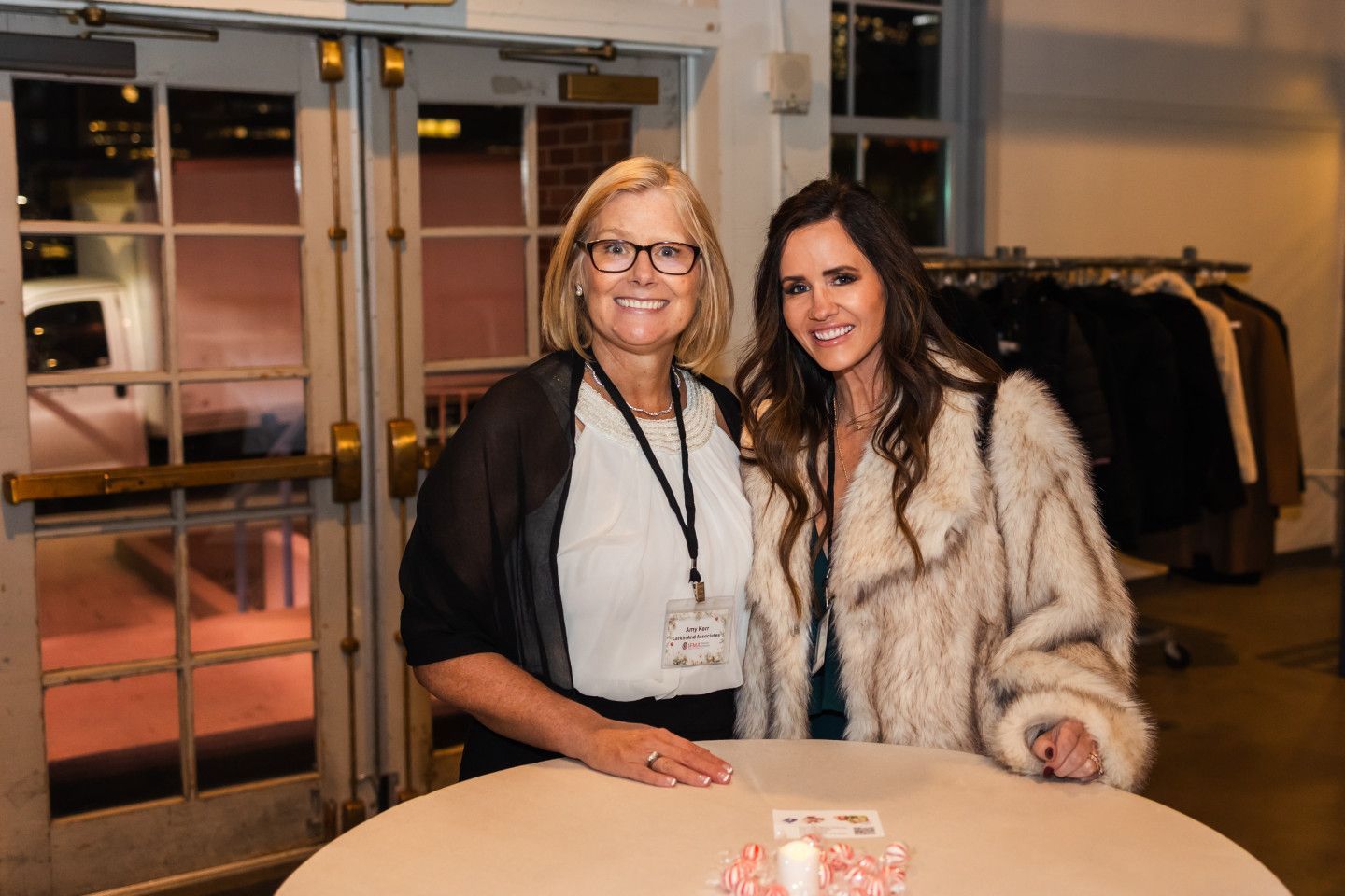Two women at an event, standing near a table. The younger woman wears a fur coat.