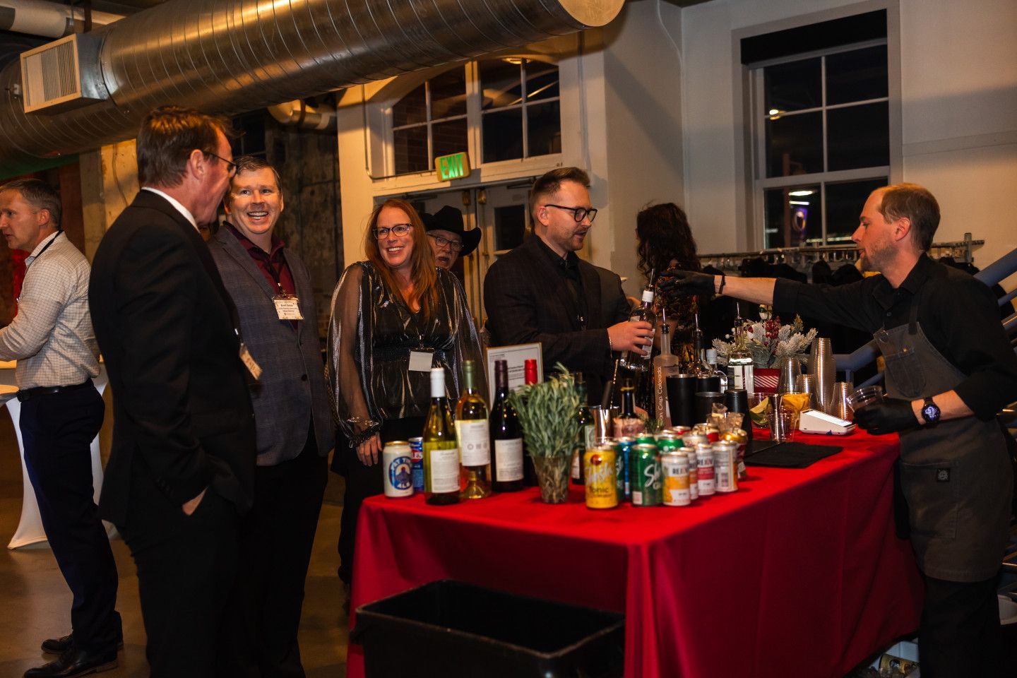 People at an event gather at a red-covered bar while a bartender serves drinks.