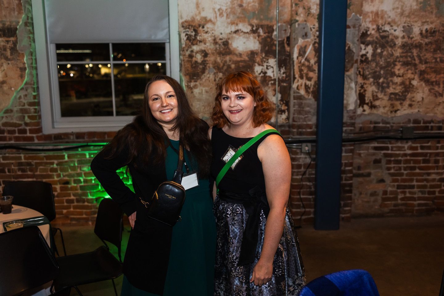 Two women smiling and posing at an event. Dark clothing, one with a sequined skirt, against a brick wall.