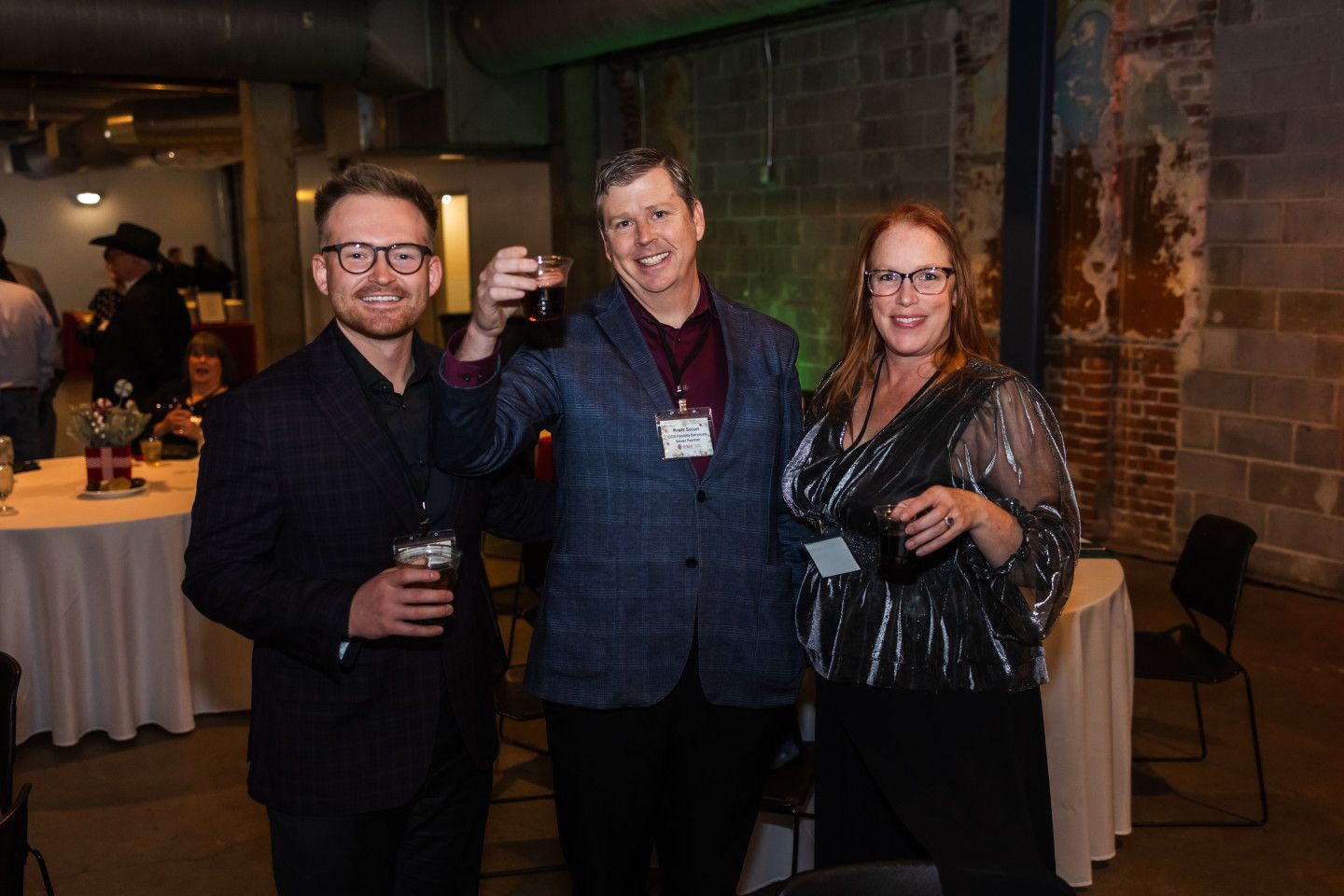 Three people at event, man raising drink. Brick wall, round tables.