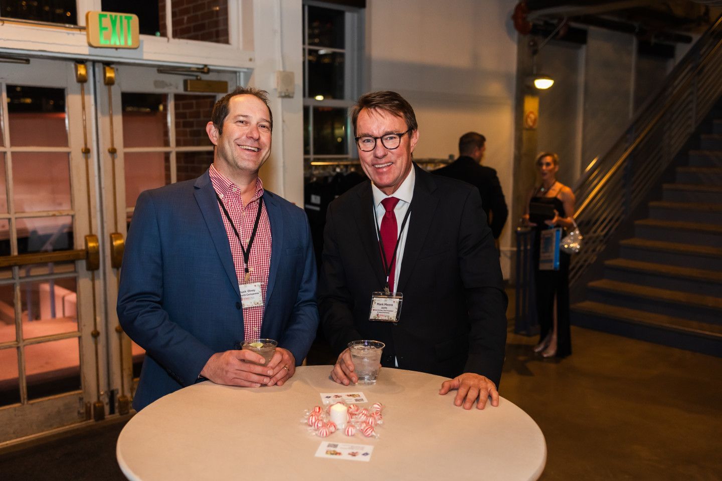 Two men at a reception, standing by a small table. One wears a blazer over a patterned shirt; the other, a suit and tie.
