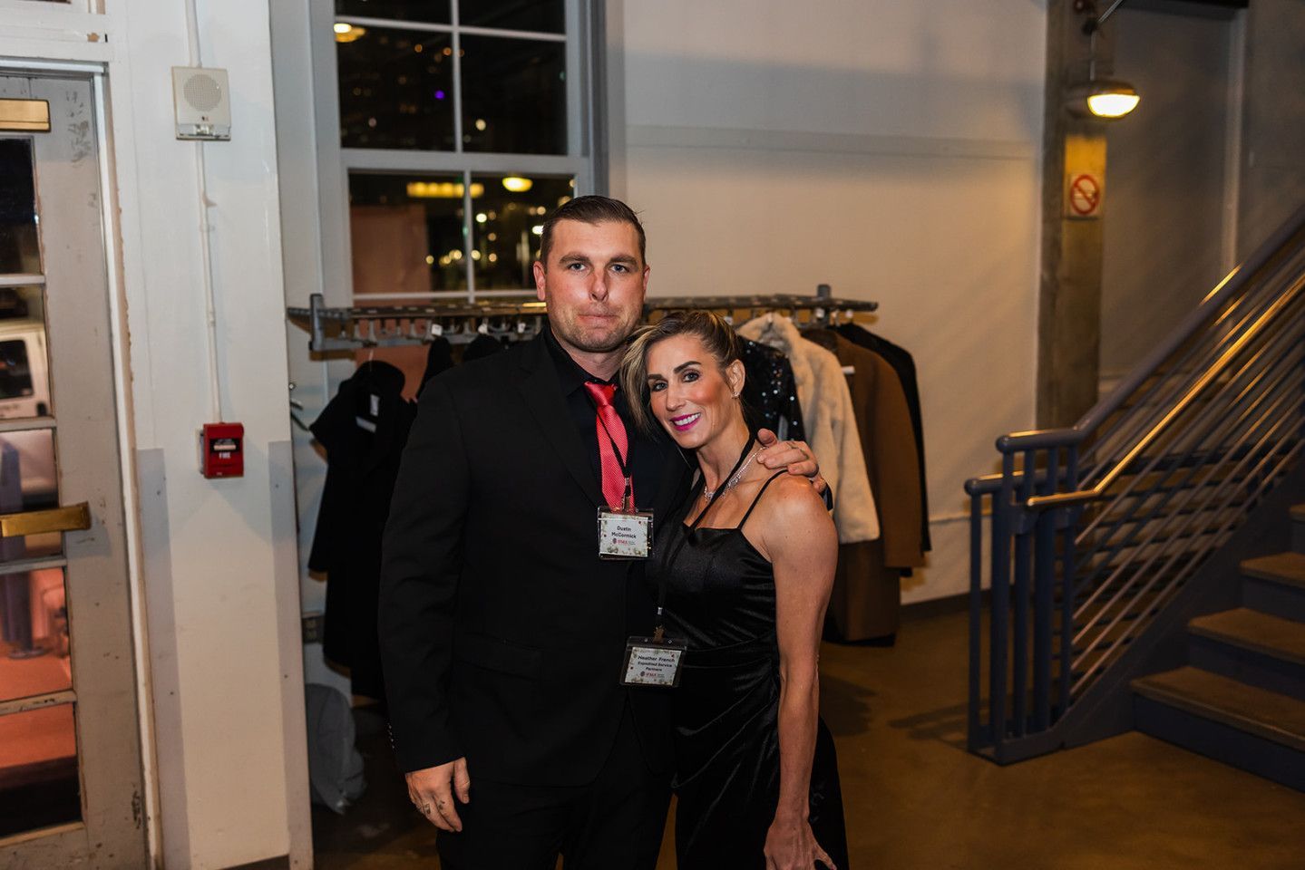 A man and woman pose indoors. The woman wears a black dress, and the man wears black with a red tie.