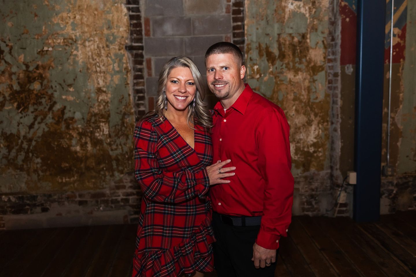 Couple posing, woman in red plaid dress, man in red shirt. Rustic interior backdrop.