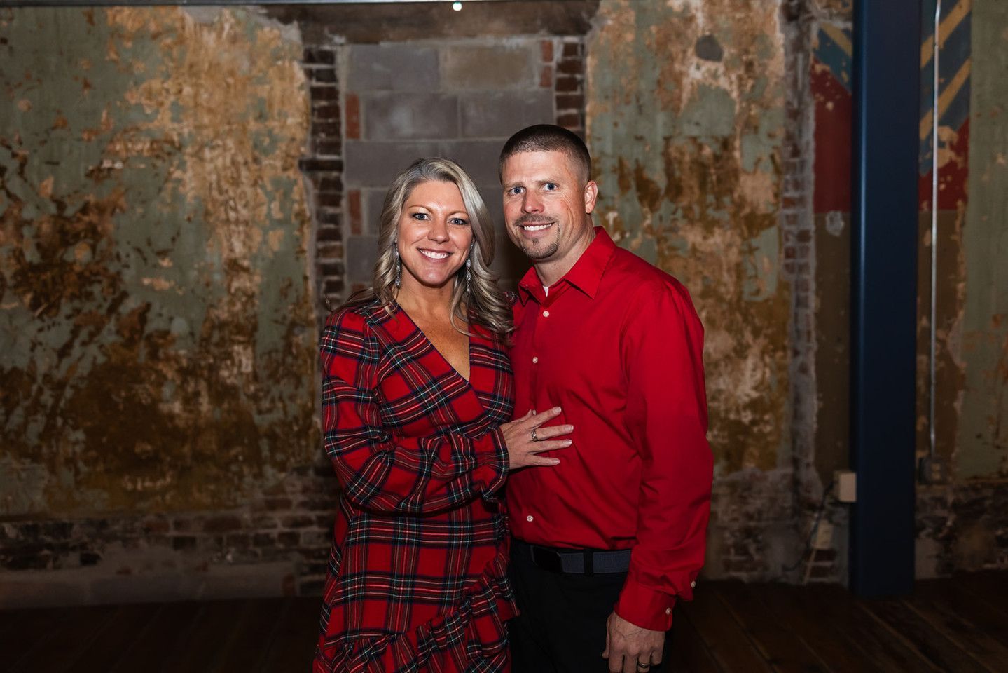 Woman in plaid dress and man in red shirt pose, smiling in front of distressed wall.