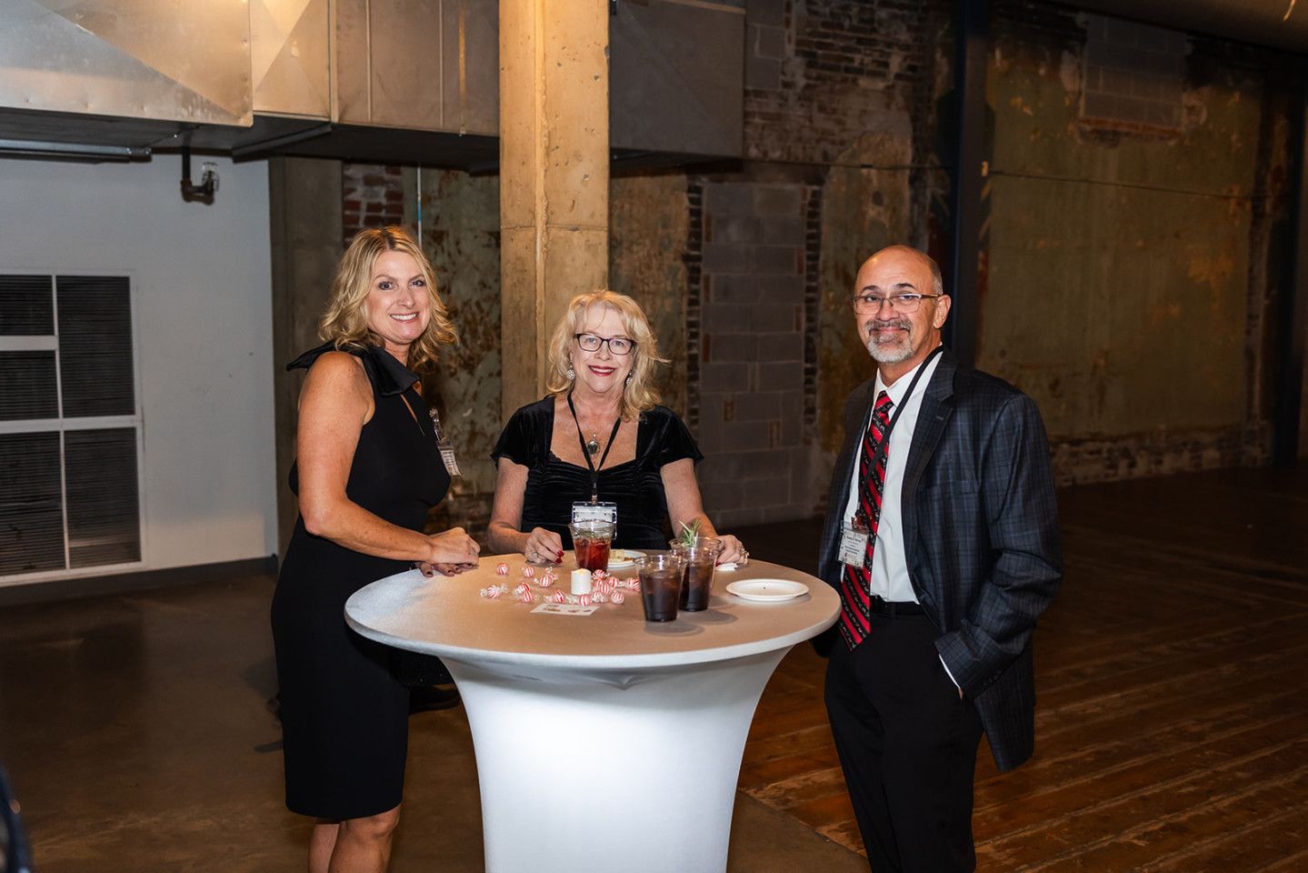 Three people at a cocktail table, in a large industrial setting. Two women and a man, smiling and looking at the camera.