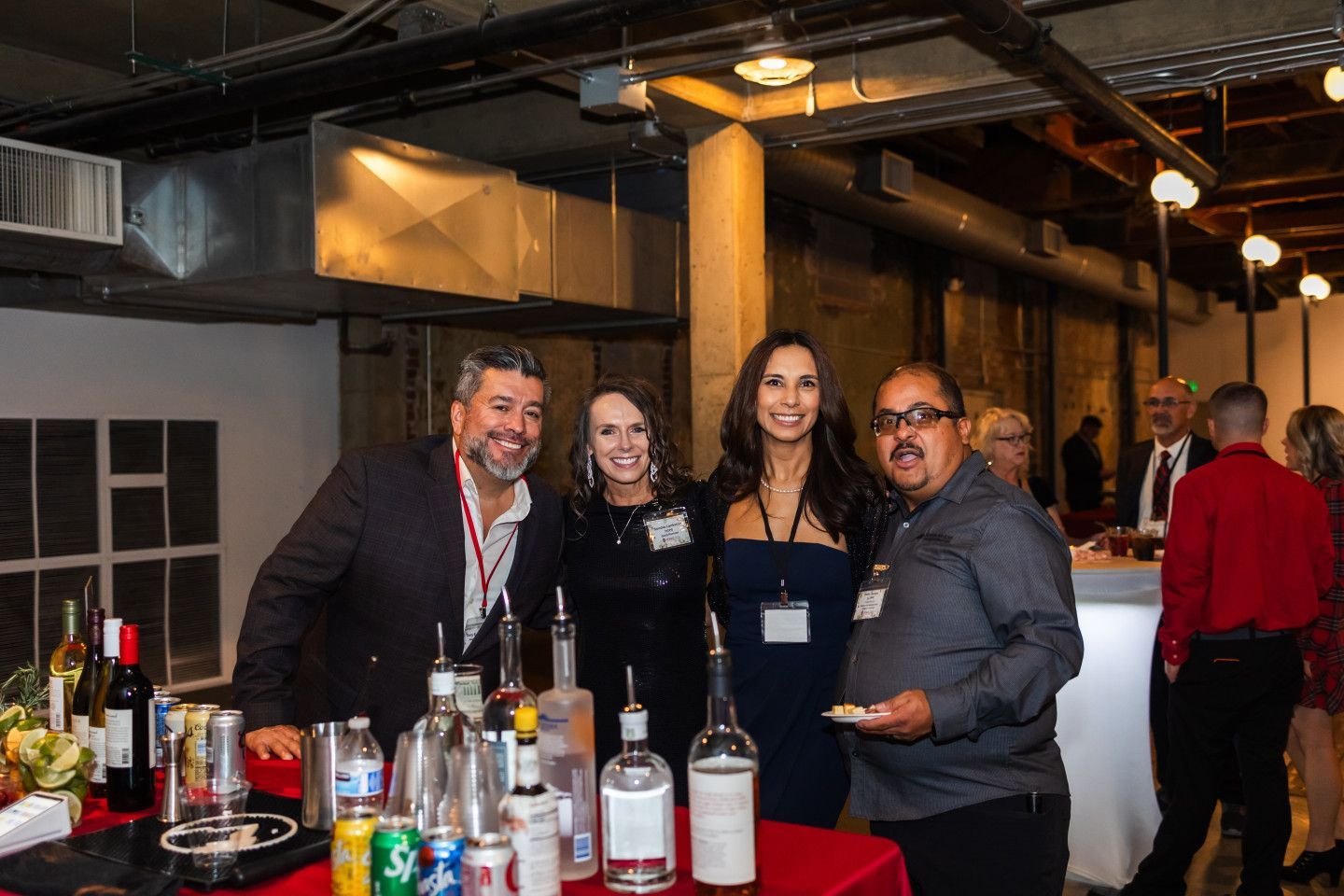 People at a bar, smiling. Bottles of alcohol on red table. Industrial-style interior.