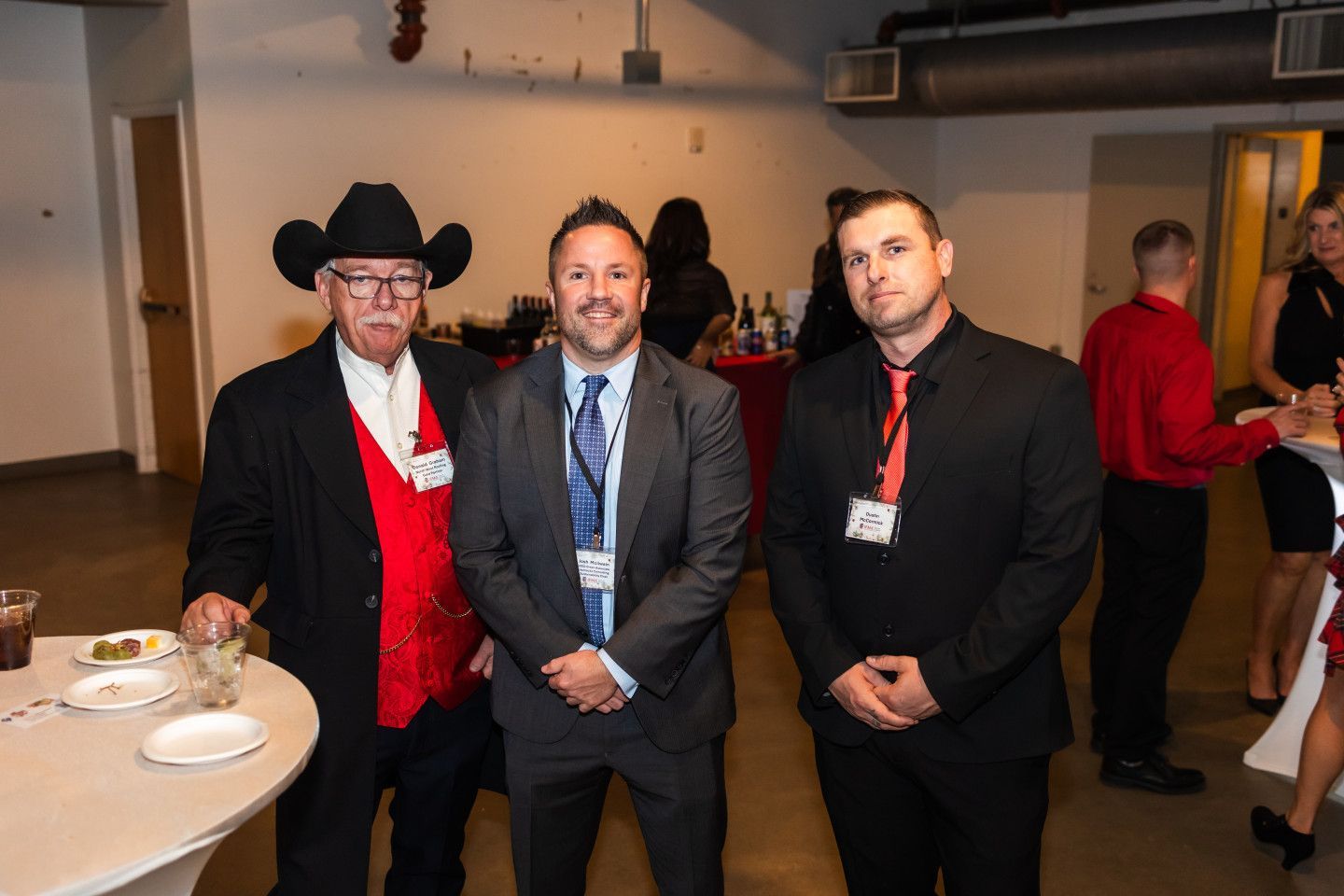 Three men at an event; one in a cowboy hat and red vest, one in a suit, and one in black with a red tie.