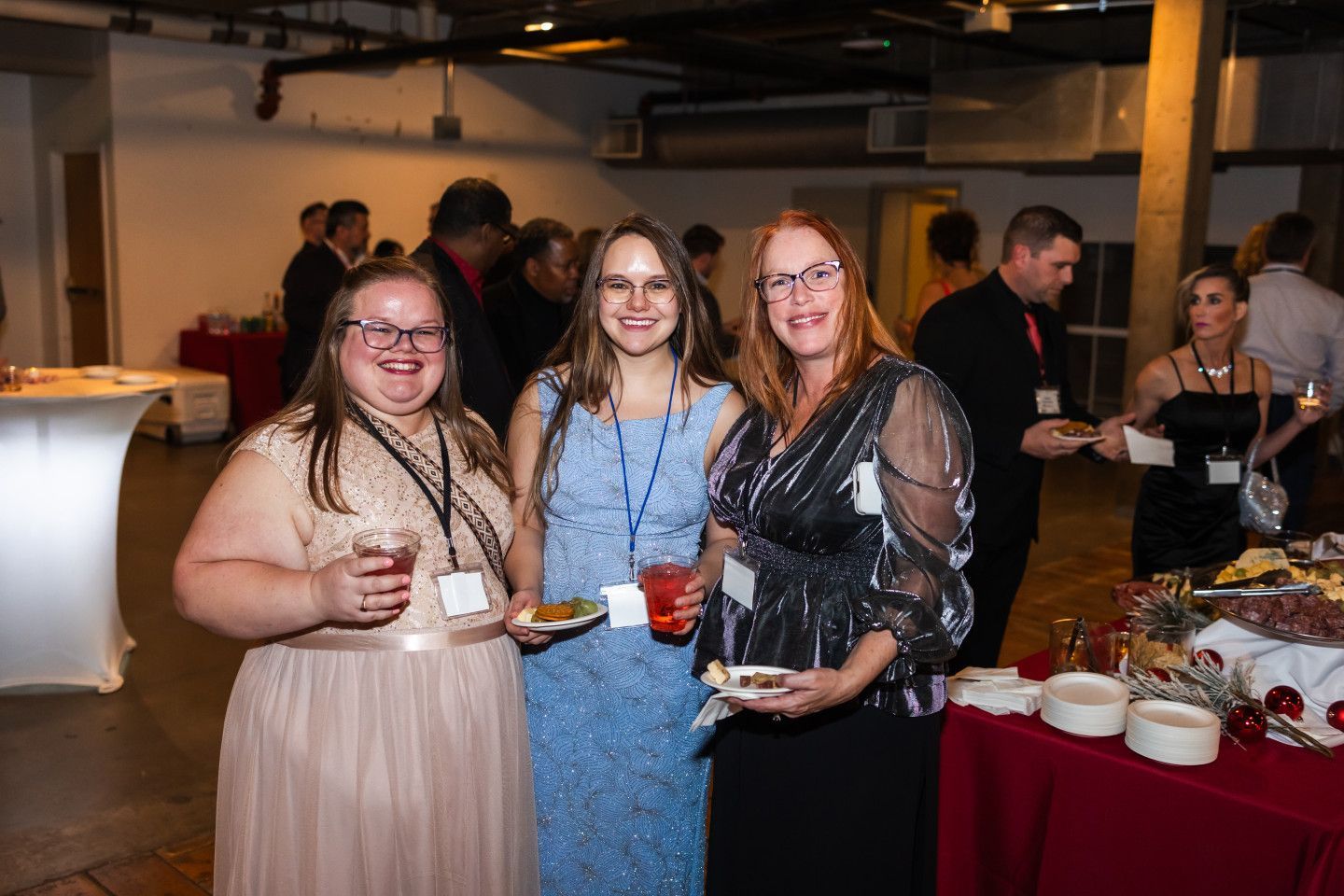 Three women smiling at a social gathering, holding drinks and food near a buffet table.