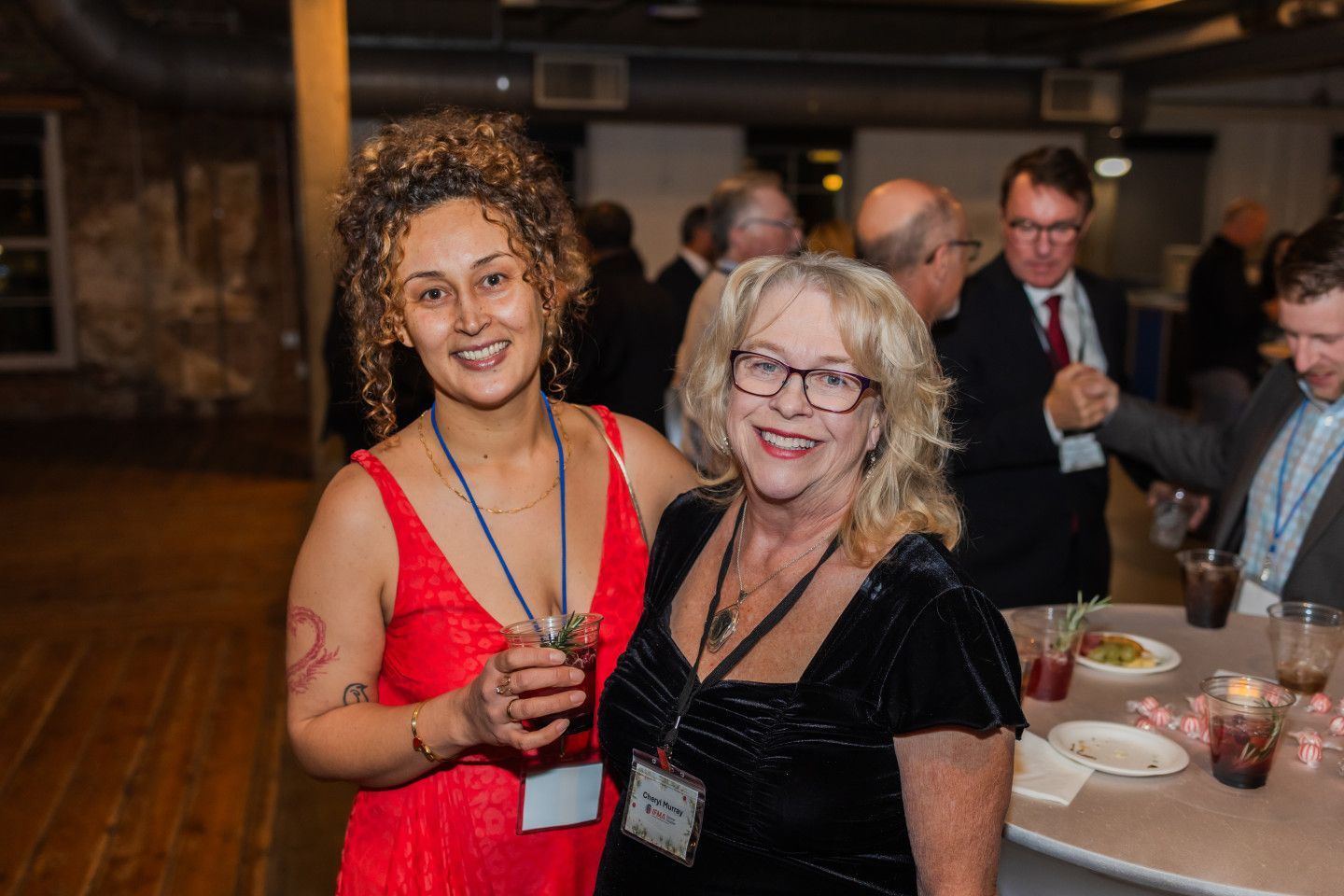Two women smiling at a gala event, one in red dress, one in black velvet. Other guests in background.