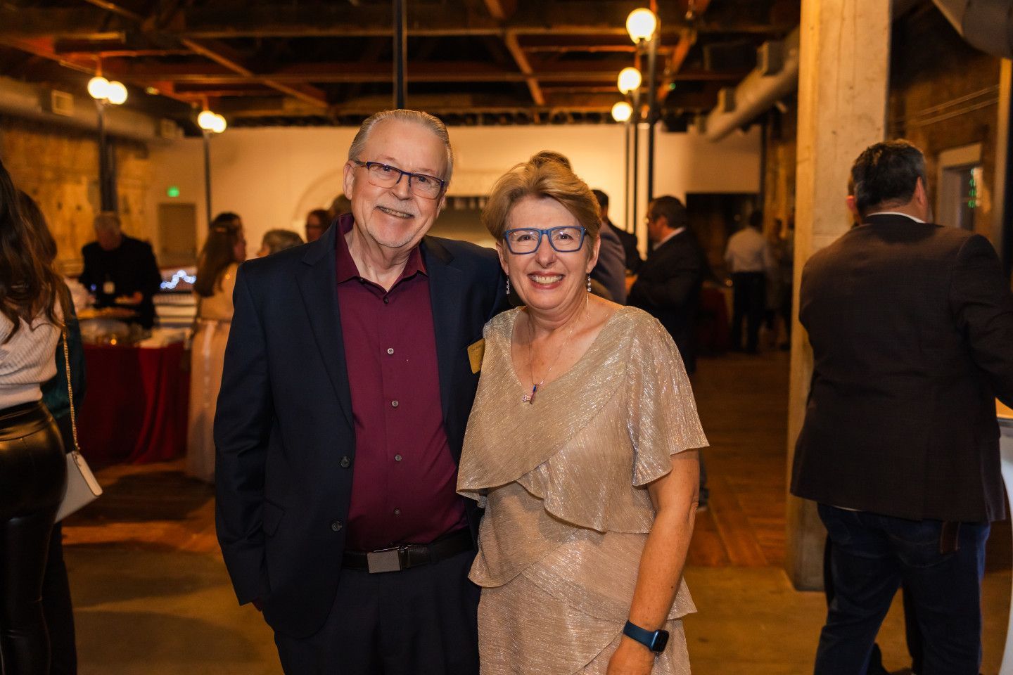 Man and woman in formal attire smile, pose together at an event with others in the background.