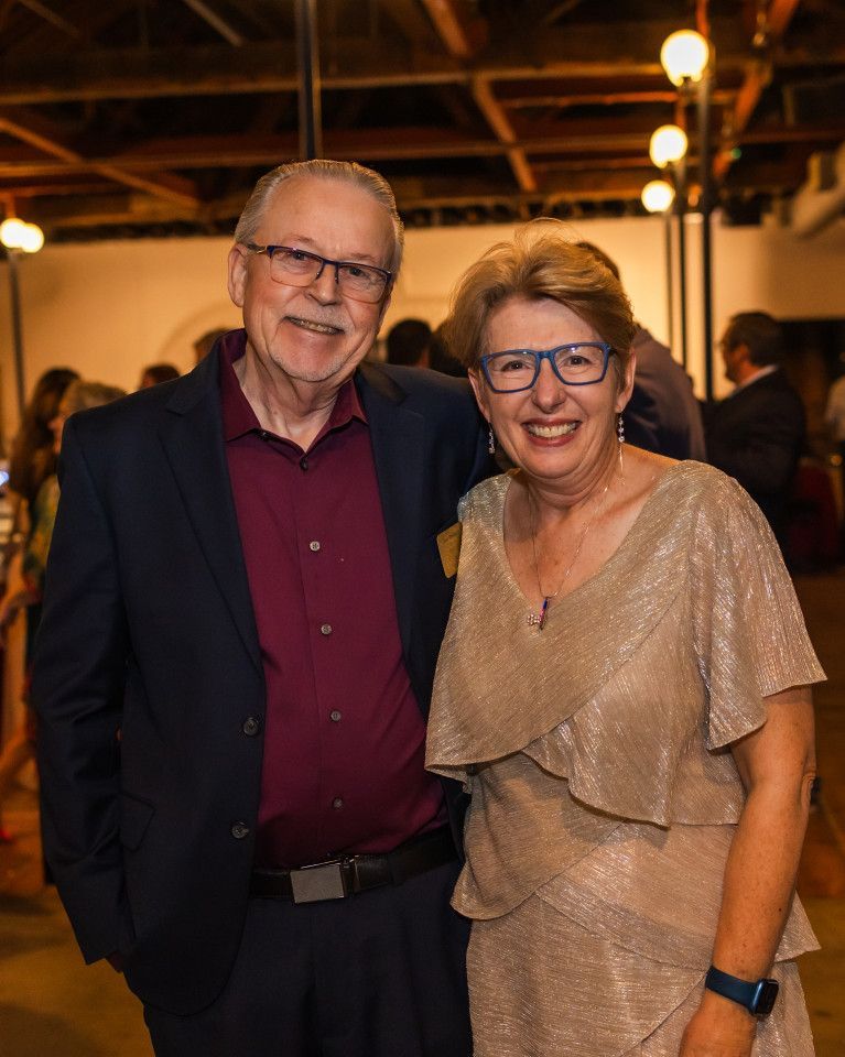 Man in suit with glasses and woman in gold dress, smiling and posing indoors.