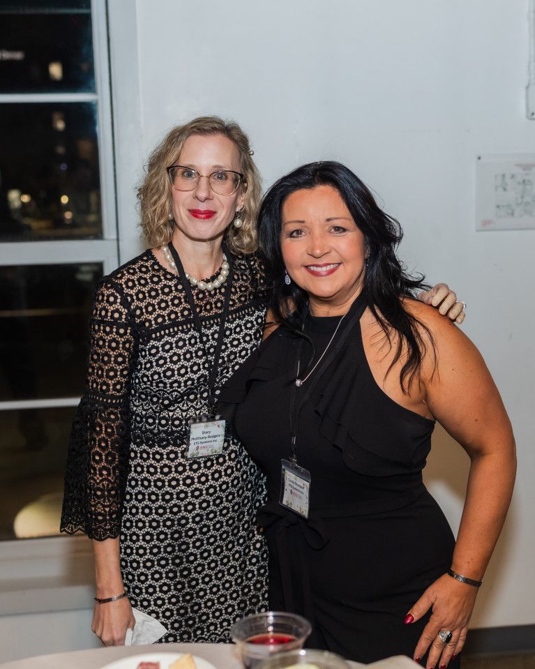Two women smiling, posing for a photo. One in a black dress, the other in a patterned dress with glasses.