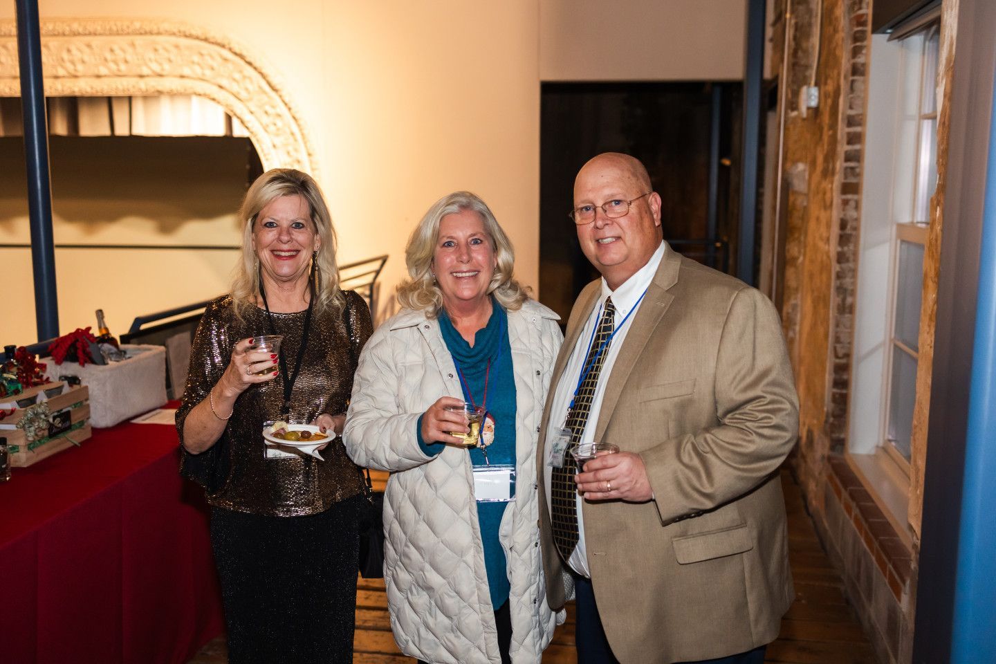 Three people at an event, holding drinks. Woman in sequin top, woman in quilted jacket, and man in blazer.