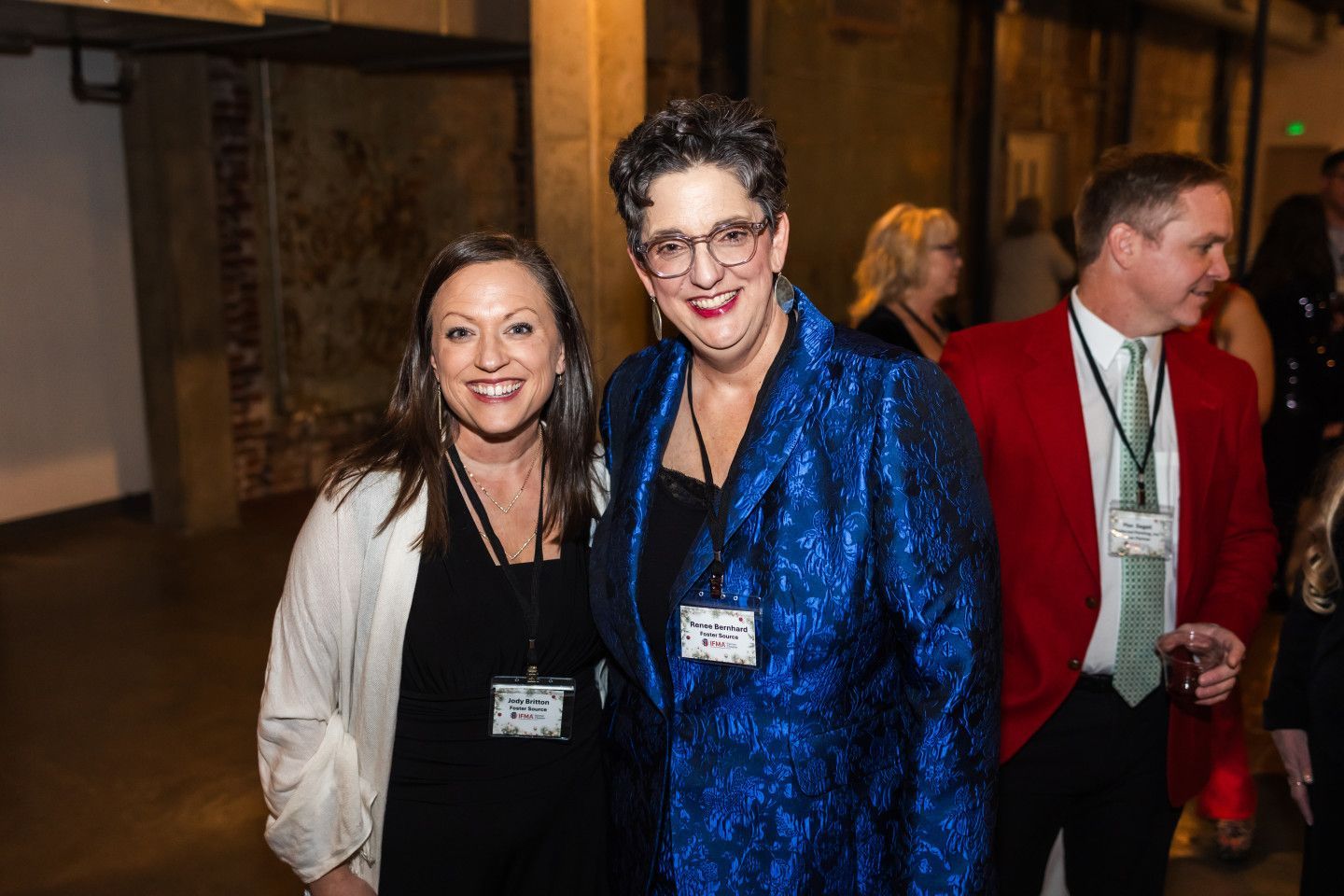 Two women smile, pose together at event. One in black, white jacket; other in blue jacket. Man in red blazer in background.