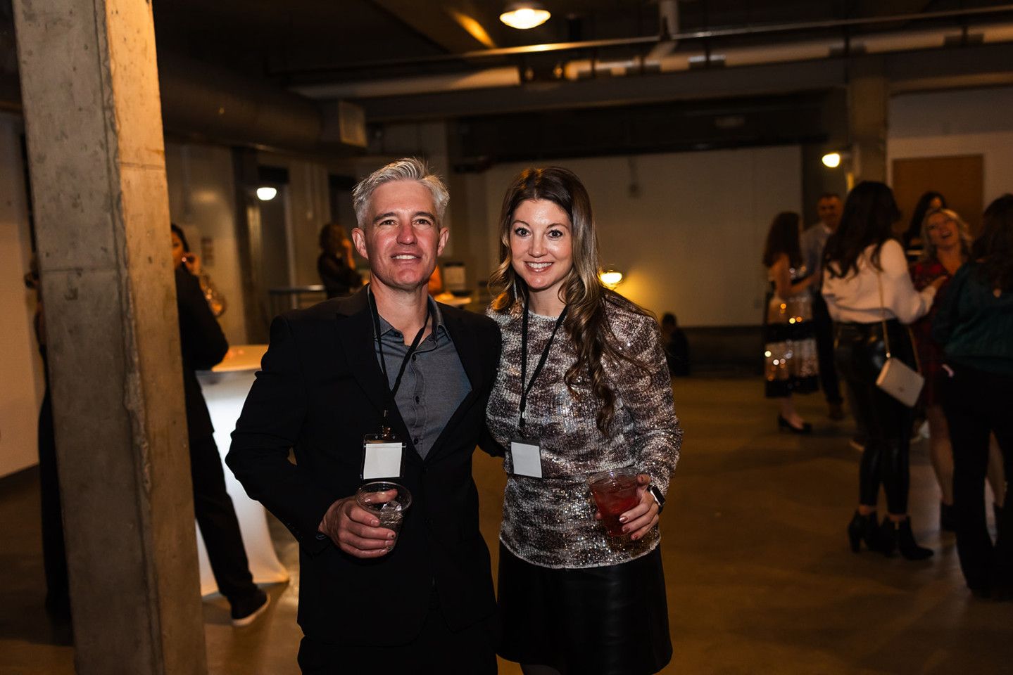 Man and woman at an event, smiling, holding drinks. Interior lit by overhead lights. Other people in background.