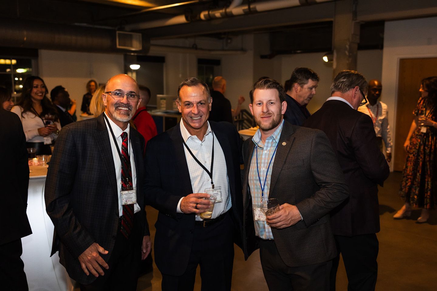 Three men smiling, holding drinks, at a gathering. One in a suit and tie, two in jackets with lanyards.