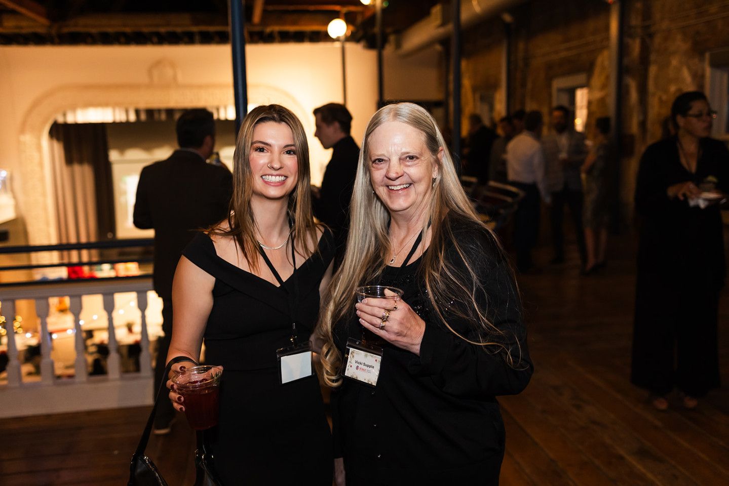 Two women at an event, smiling, wearing black dresses and name tags, holding drinks.