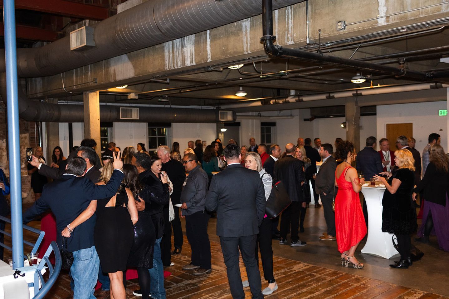 Party in a spacious room. Guests mingle, some with drinks. Exposed pipes and beams overhead, wood floor.