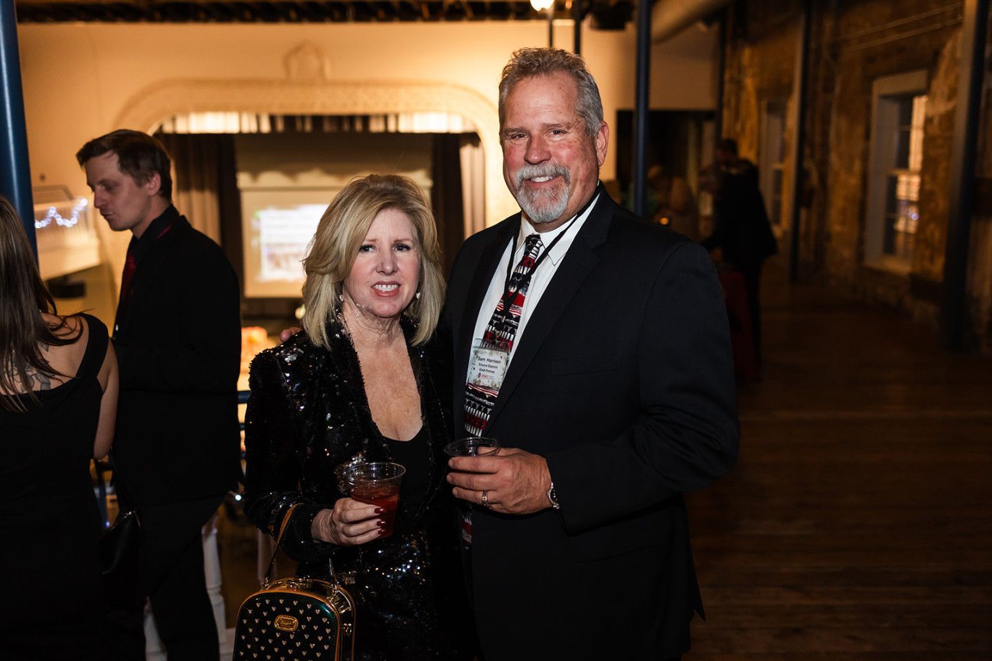 A couple smiles, holding drinks at a formal event, with dark clothing and a blurred background.