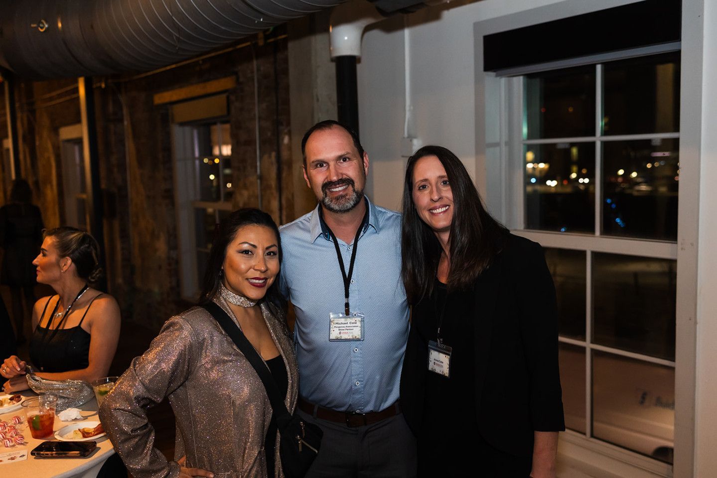 Three people smiling, posing together at an event. Indoors, near a window, with a partially visible background.