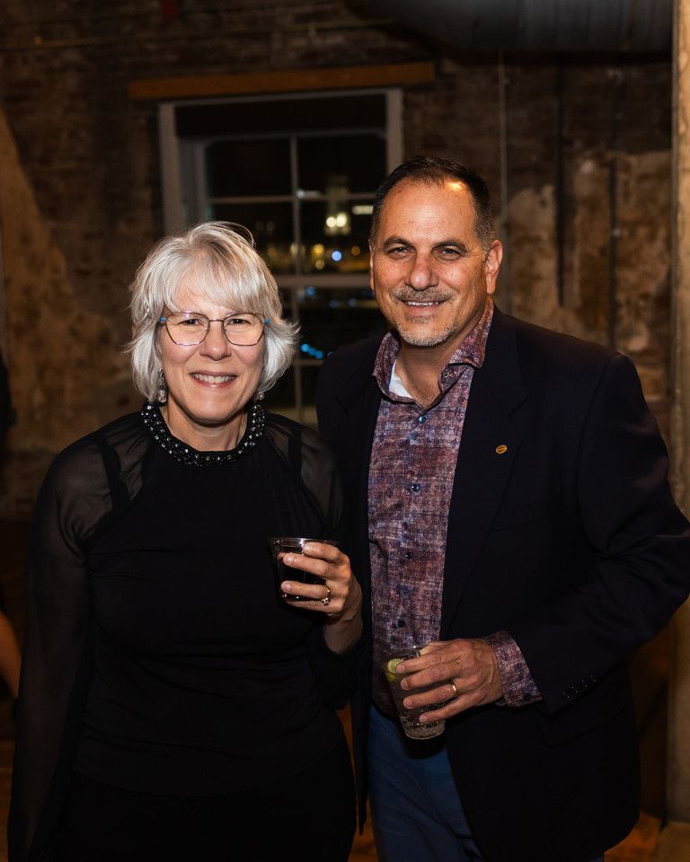 Woman with glasses and man in blazer pose together indoors, smiling.