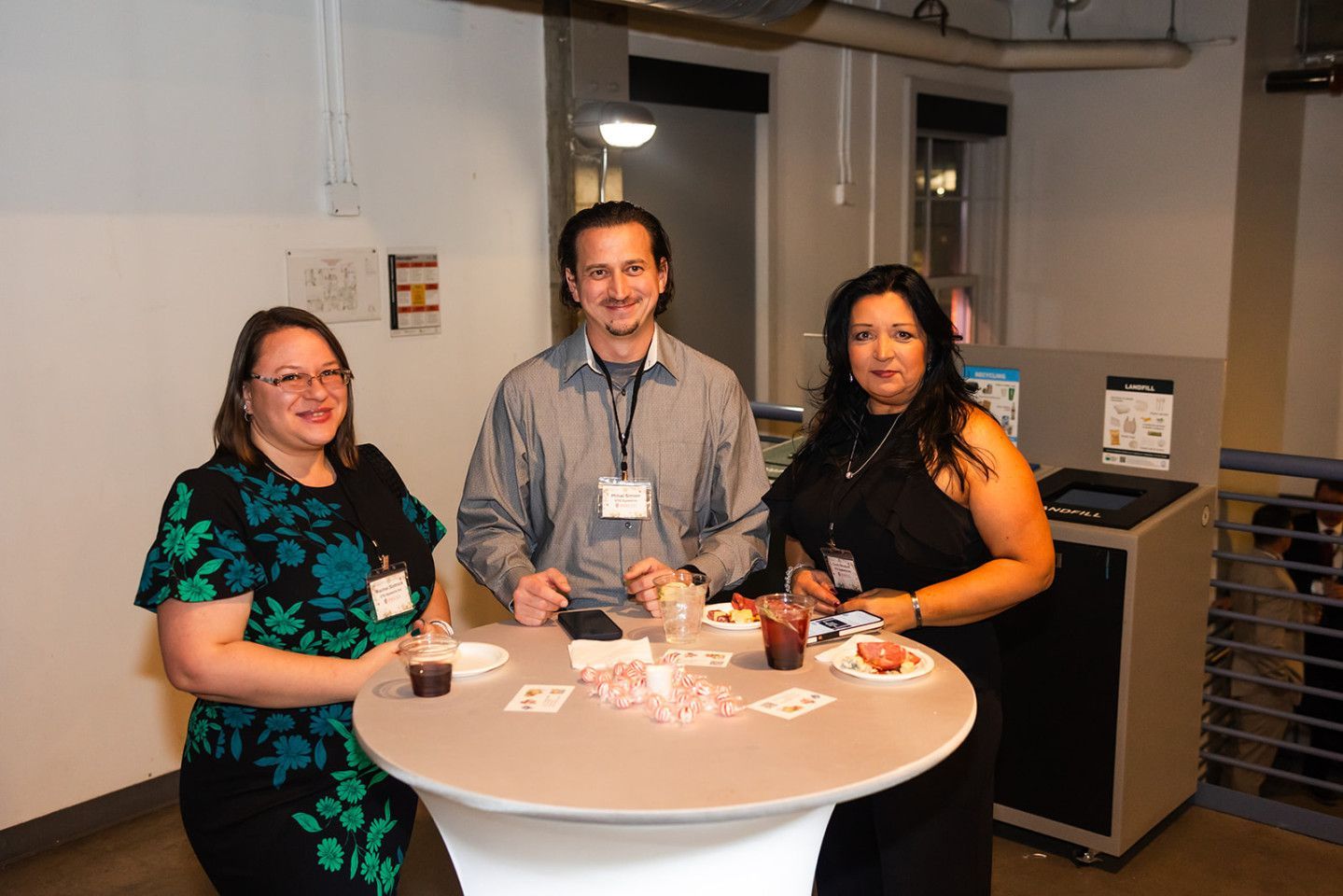 Three people at a table with drinks and snacks. Indoor setting, white walls, and a small table.