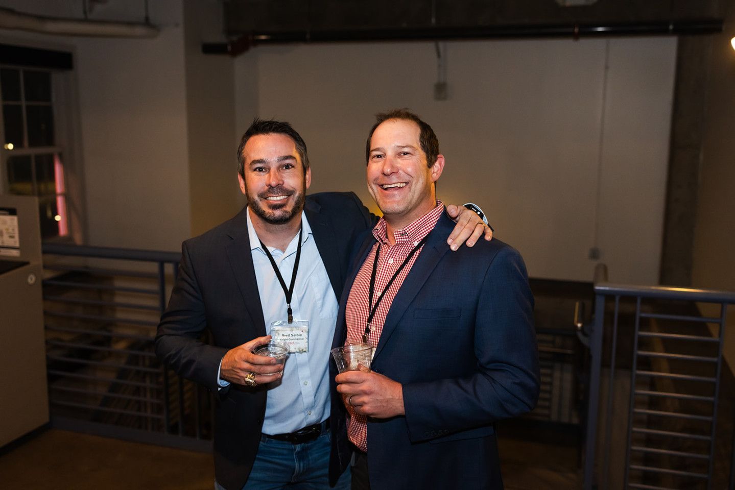 Two men smiling, arm around each other, holding drinks at an event.